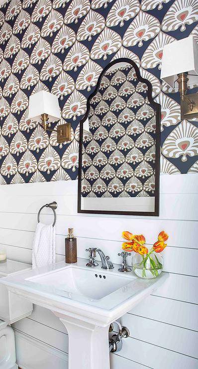 Between brass sconces, a black arch mirror is hung from a wall clad in taupe and blue wallpaper lined with shiplap trim. The mirror is mounted over a white pedestal sink finished with a polished nickel cross handle faucet kit.