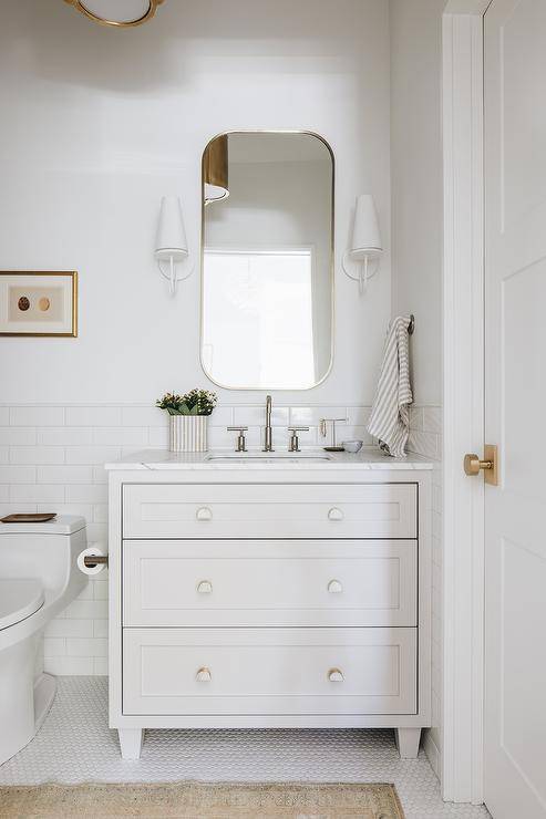 Aerin Fontaine Single Sconce flank a curved white mirror hung over an off-white dresser-like washstand finished with a polished nickel gooseneck faucet and white and gold half moon knobs. The washstand is mounted in front of a white subway backsplash tiles.
