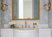 Romantic French-style bathroom is lit by facing gold candle sconces mounted on either side of a gold rope mirror hung from a wall covered in blue and white floral wallpaper. The mirror is mounted above a white half-moon French style sink vanity paired with a polished brass hook and spout faucet.