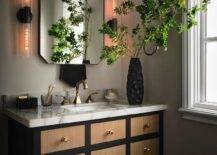 Bathroom features a brown and black apothecary style washstand with brass knobs and a marble countertop with brass faucet under a gold and black mirror flanked by long sconces.