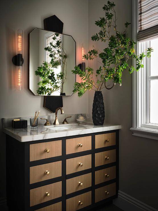 Bathroom features a brown and black apothecary style washstand with brass knobs and a marble countertop with brass faucet under a gold and black mirror flanked by long sconces.