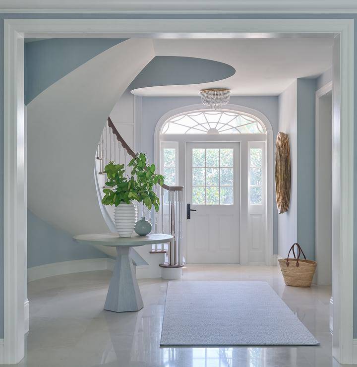 Morgan Harrison Home - A white front door with sidelights fixed beneath an arched transom window opens to a white and blue entry lit by an Aerin Jacqueline Flush Mount. A round oak table topped with white and gray vases and placed beside a curved staircase boasting a brown wood handrail on white spindles.