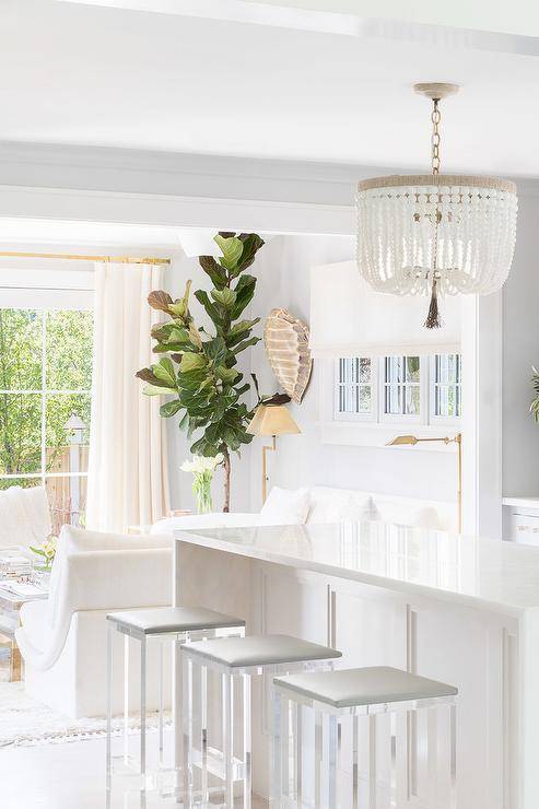 displaying a stunning white beaded chandelier over a white waterfall quartz island paired with lucite backless counter stools topped with a light gray cushion. White furnishings in the adjacent room and cream curtains are balanced by green homey house plants and gold fixtures to create a light, airy and fresh living space.