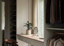 A white floating makeup vanity with a wooden countertop is fixed in a walk-in closet in front of a window and between stacked clothing rails and built-in gray shoe shelves.