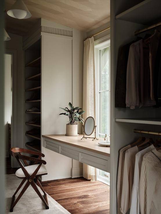 A white floating makeup vanity with a wooden countertop is fixed in a walk-in closet in front of a window and between stacked clothing rails and built-in gray shoe shelves.