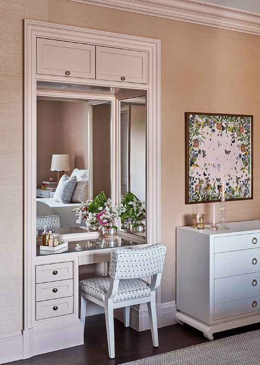 A white and blue vanity chair sits at pink makeup vanity with a mirrored top built in a nook and finished with pink framed vanity mirrors.
