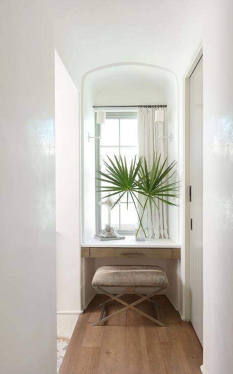 A cowhide bench sits in a makeup vanity nook at a brown floating vanity positioned under a window dressed in short white curtains.