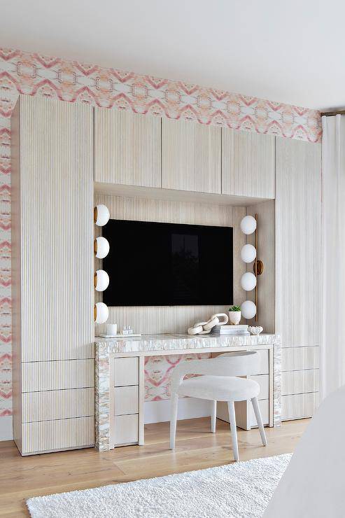 A white sleek vanity chair sits at a pink and gray marble waterfall edge makeup vanity fixed against a reeded backsplash beneath a flat panel TV hung beneath and between reeded cabinets. The vanity is lit by facing brass and white glass 4-light vanity light.