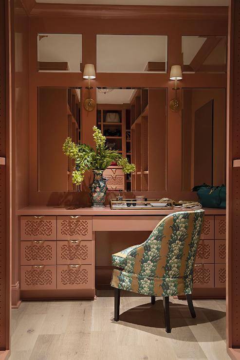 Paneled mirrors are lit by brass sconces fixed to a pink wall over a pink makeup vanity accented with lattice drawers fronts and brass pulls. The vanity is paired with a green and brown chair.