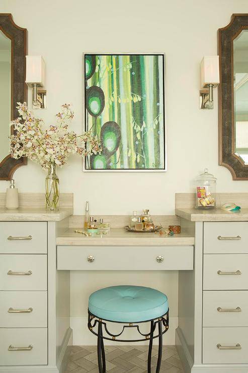A green art piece hangs over a light gray drop-down makeup vanity fixed between light gray washstands and topped with a taupe stone countertop seating a turquoise stool.