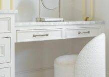 A white boucle chair sits on a white staggered floor tiles at a white drop-down makeup vanity finished with a marble countertop fixed under stacked art.