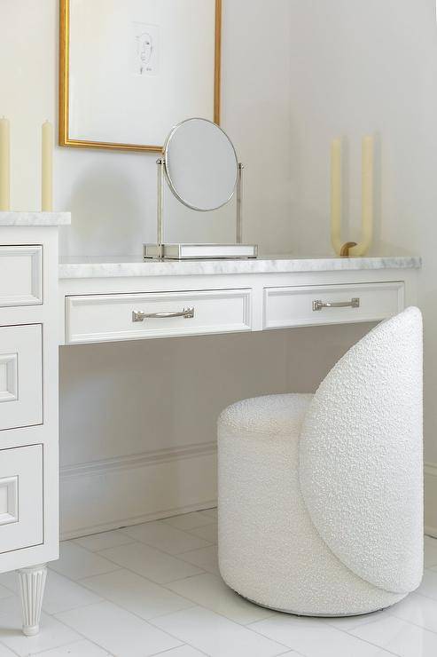 A white boucle chair sits on a white staggered floor tiles at a white drop-down makeup vanity finished with a marble countertop fixed under stacked art.