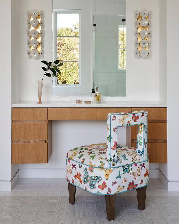 A butterfly print stool sits at a brown veneer floating makeup vanity fixed under a frameless vanity mirror lit by white and gold sconces.