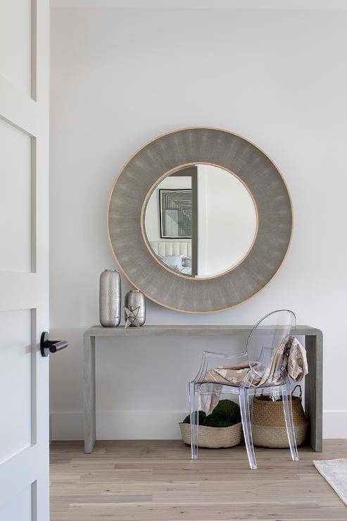 A ghost chair sits at a gray waterfall makeup vanity positioned beneath a round gold and gray shagreen mirror.