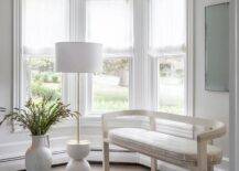 Living room features a light taupe curved bench with a white and taupe cushion lit by a white and brass floor lamp with tall potted plant.