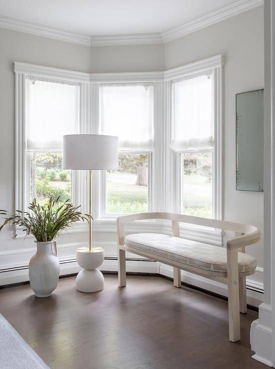 Living room features a light taupe curved bench with a white and taupe cushion lit by a white and brass floor lamp with tall potted plant.