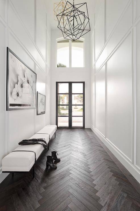 Galley style two story foyer with dark stained wood herringbone floors features a black and white bench under black and white floral art, white wall moldings and a black frame glass panel front door.