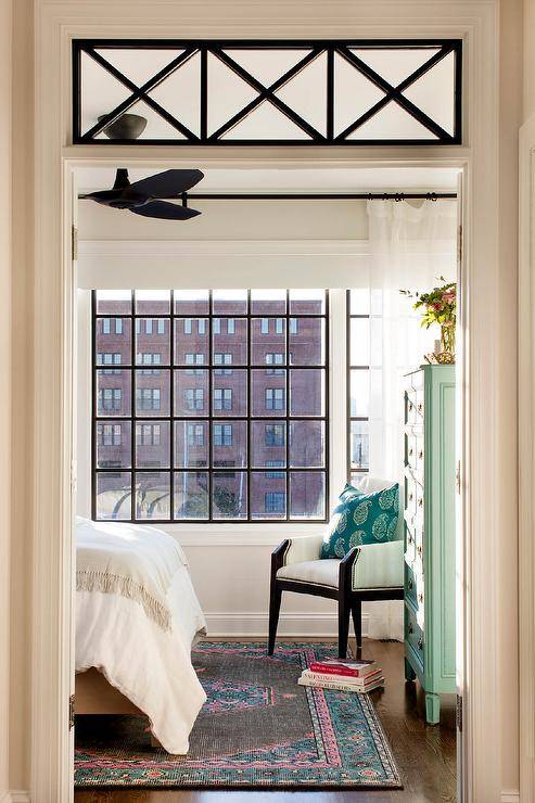 Bedroom with transom window inviting natural light highlighting vintage style decor and furnishings. A black and white chair is furnished in a corner beside a green tall boy vintage dresser with ring pulls finished with a pink and blue area rug under a bed.