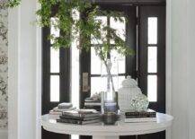 Flanked by sidelights and positioned under a transom window, a black front door with glass panels opens to a foyer boasting a round white styled entry table lit by a flush mount lantern.