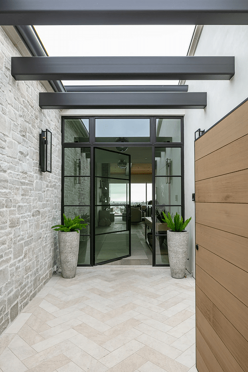 Cream travertine herringbone pattern pavers lead to a steel and glass front door flanked by sidelight and positioned beneath a transom window.