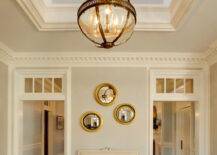 Incredible two story foyer illuminated by a Restoration Hardware Victorian Hotel Pendant over gray walls finished with dentil crown molding above doorways with transom windows. The foyer features an ivory French settee layered with light gray pillows below three gold convex mirrors over hardwood floors topped with a bound diamond sisal rug.