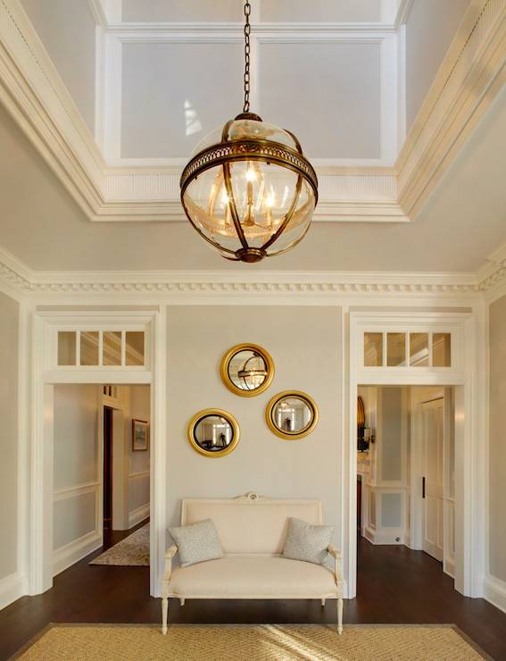 Incredible two story foyer illuminated by a Restoration Hardware Victorian Hotel Pendant over gray walls finished with dentil crown molding above doorways with transom windows. The foyer features an ivory French settee layered with light gray pillows below three gold convex mirrors over hardwood floors topped with a bound diamond sisal rug.