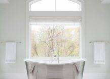 Restoration Hardware Victorian Hotel Pendant illuminates a master bathroom above a silver modern cast iron bathtub designed at a picture window fitted with a white Roman shade. White floor tiles lighten the bathroom with a sophisticated appeal accented with white mosaic carpet tiles.