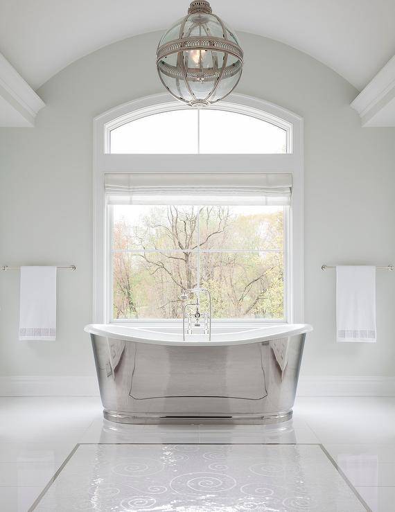 Restoration Hardware Victorian Hotel Pendant illuminates a master bathroom above a silver modern cast iron bathtub designed at a picture window fitted with a white Roman shade. White floor tiles lighten the bathroom with a sophisticated appeal accented with white mosaic carpet tiles.