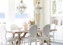 White French round back chairs sit on a white rug around a trestle dining table lit by Restoration Hardware Victorian Hotel Pendants hung from a coffered ceiling. At the end of the table, white built in cabinets are located beneath glass front cabinets finished with a white x-trim.