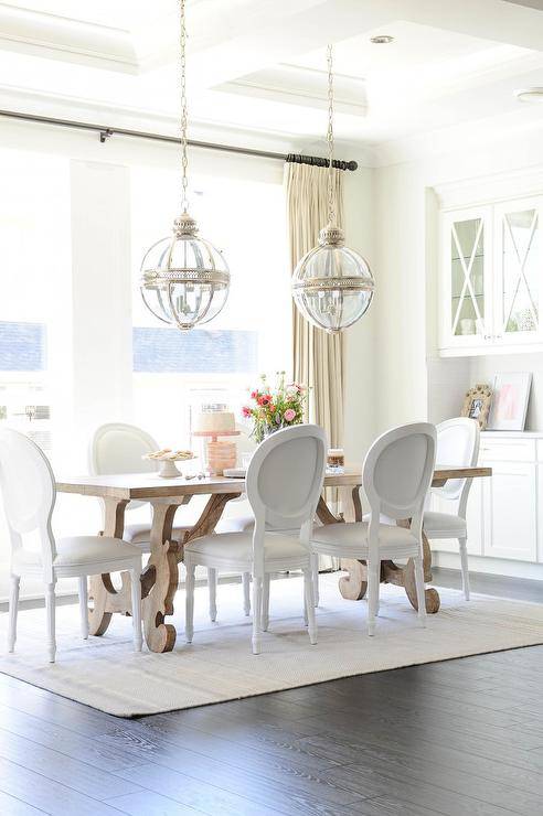 White French round back chairs sit on a white rug around a trestle dining table lit by Restoration Hardware Victorian Hotel Pendants hung from a coffered ceiling. At the end of the table, white built in cabinets are located beneath glass front cabinets finished with a white x-trim.