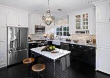 L shaped kitchen features white upper cabinets and black lower cabinets paired with marble countertops and a marble subway tiled backsplash. A small farmhouse sink and a vintage gooseneck faucet stands adjacent to a microwave hood and stainless steel stove. A Restoration Hardware Victorian Hotel Pendant illuminates an iron and marble kitchen island, Crate & Barrel French Kitchen Island, lined with two industrial swivel bar stools placed atop an ebony floor.