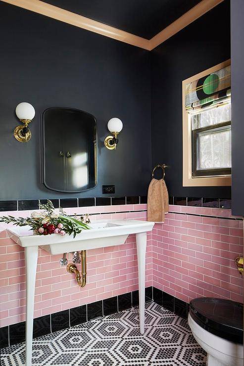 Gold and black sconces are mounted to a black painted wall on either side of a curved vanity mirror hung above pink offset wall tiles finished with black border tiles. A white French sink vanity is fixed in front of the backsplash tiles and against black and white hexagon floor tiles.