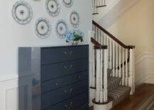 Elegant hallway with raised panel wainscoting below pale blue upper walls accented with blue wall plates hung above a dark blue lacquered chest of drawers atop hardwood floors layered with a blue and brown striped rug runner which continues up the stairway.