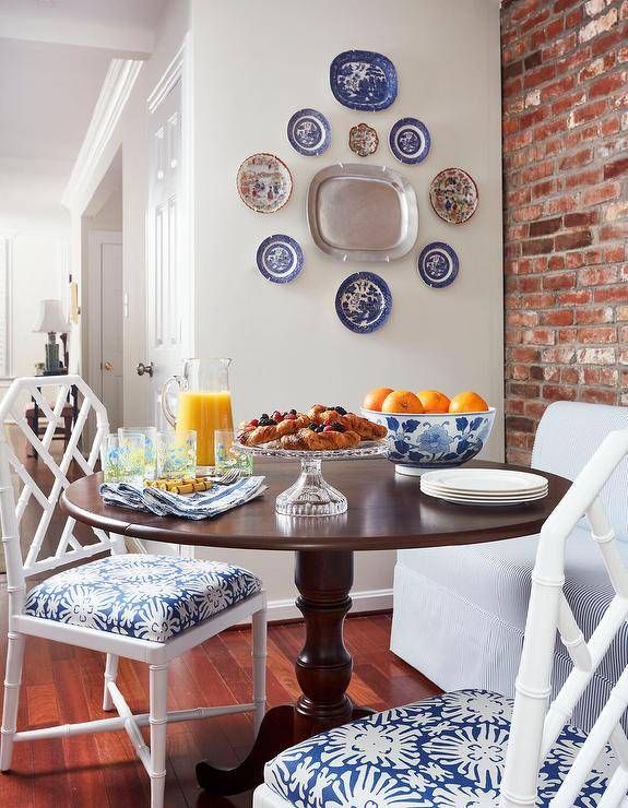 Dining space features white bamboo chairs with white and blue cushions and a blue pinstripe dining bench at a round traditional dining table with a brick accent wall and a decorative plate gallery.