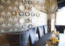 Incredible dining room with F. Schumacher Tashkent Ikat Silver wallpaper and Fornasetti Plate wall decor. Amazing art deco gold chandelier over the chic black dining table with gray upholstered dining chairs with nailhead trim. Dining table filled with a collection of gold and white bowls.