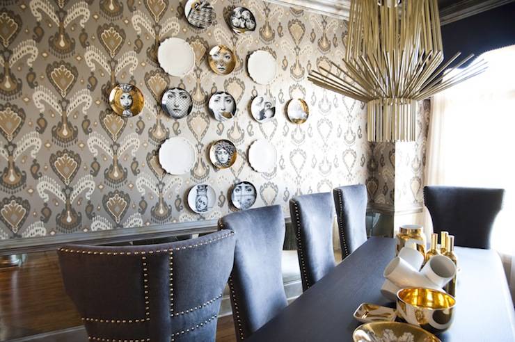 Incredible dining room with F. Schumacher Tashkent Ikat Silver wallpaper and Fornasetti Plate wall decor. Amazing art deco gold chandelier over the chic black dining table with gray upholstered dining chairs with nailhead trim. Dining table filled with a collection of gold and white bowls.
