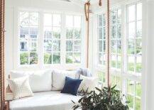 Hung in a gorgeous sunroom from a vaulted plank ceiling, a wood and rope swing sofa is accented with white cushions and hangs over gray quatrefoil floor tiles facing a round woven ottoman.