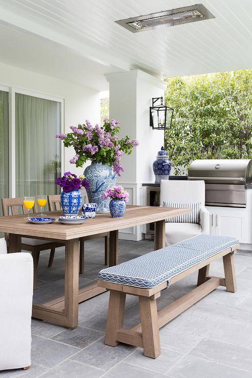 Brown teak dining table and a matching dining bench feature a blue and white pattern upholstered cushion. Natural linen slipcovered dining chairs mix with the bench and teak chairs atop slate pavers next to a covered patio bbq.