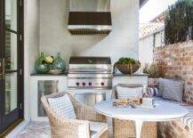 Outdoor patio designed with a red brick bench and wall upholstered with a gray and white striped cushion. A round outdoor dining table is paired with a set of wicker chairs next to a cooktop designed with a small wolf bbq, stainless steel vent hood, and kitchen space.