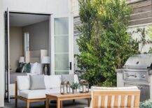 A teak chair and a teak sofa accented with white cushions and gray pillows sit on either side of a teak coffee table positioned to the side of a BBQ.