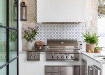 Well appointed l-shaped outdoor kitchen boasts a white hood fixed to gray and blue mosaic concrete backsplash tiles over a stainless steel BBQ.