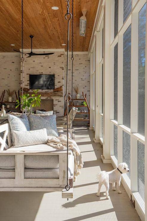 Welcoming sunroom features a whitewashed wooden swing sofa topped with pale blue pillows and hung from a brown wood plank ceiling.