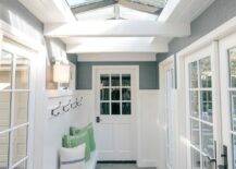 A vaulted ceiling fitted with skylights allows natural light to stream into to a sunroom boasting slate floor tiles and a white built-in bench mounted beneath a row of iron hooks fixed to a shiplap backsplash lined with a gray upper wall.