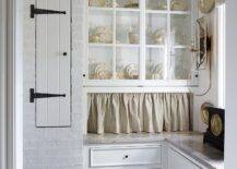 Wonderfully appointed French country kitchen pantry features glass cabinets finished with a gray skirt and positioned above white cabinets donning small oil rubbed bronze knobs.