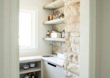 Small u-shaped kitchen pantry features an brown vintage rug placed on a dark stained wood floor in front of white shelves fixed beneath a window and adjacent to white drawers. A small stainless steel sink with a polished nickel gooseneck faucet is positioned in a corner under stacked oak floating shelves mounted against a whitewashed brick wall.