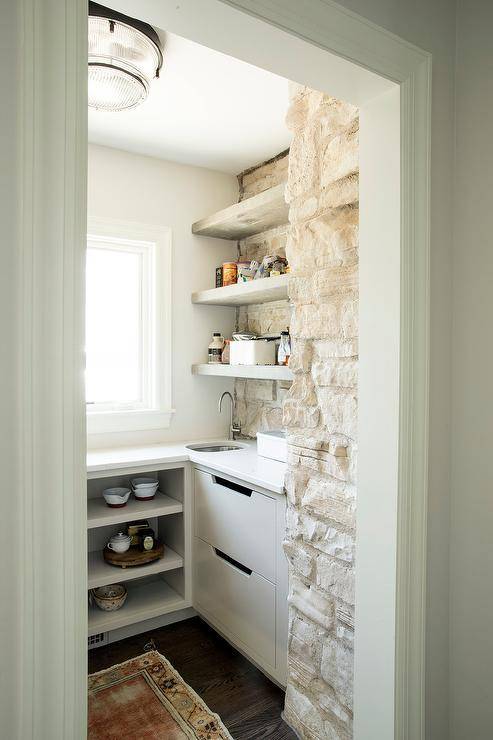 Small u-shaped kitchen pantry features an brown vintage rug placed on a dark stained wood floor in front of white shelves fixed beneath a window and adjacent to white drawers. A small stainless steel sink with a polished nickel gooseneck faucet is positioned in a corner under stacked oak floating shelves mounted against a whitewashed brick wall.