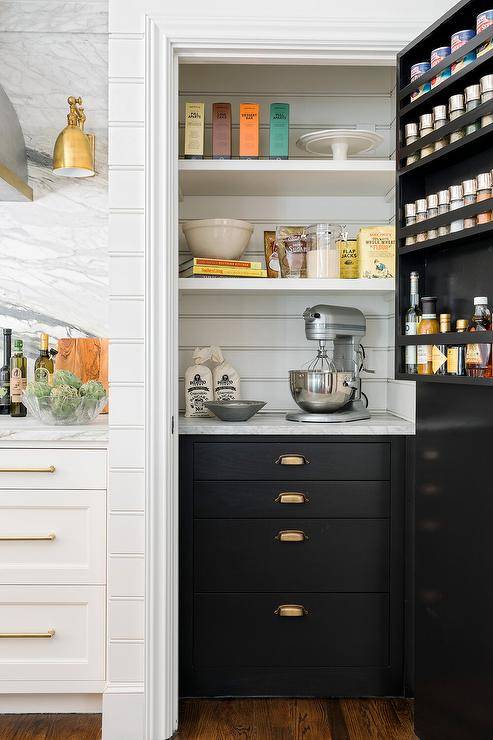 Stacked spice shelves adorn the back of a black pantry door. A black and white kitchen pantry features black cabinetry adorned with antique brass cup pulls. paired with horizontal plank backsplash.