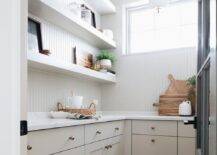 Cottage style gray walk-in pantry features gray floating shelves fixed against a gray beadboard wall over gray cabinets topped with a white quartz countertop.