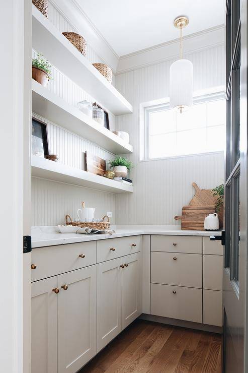 Cottage style gray walk-in pantry features gray floating shelves fixed against a gray beadboard wall over gray cabinets topped with a white quartz countertop.