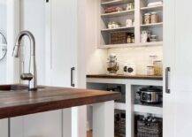 Dual white doors on oil rubbed bronze rails open to a small walk-in kitchen pantry boasting white shelves positioned above a wood countertop mounted over shelves and against white staggered wall tiles.